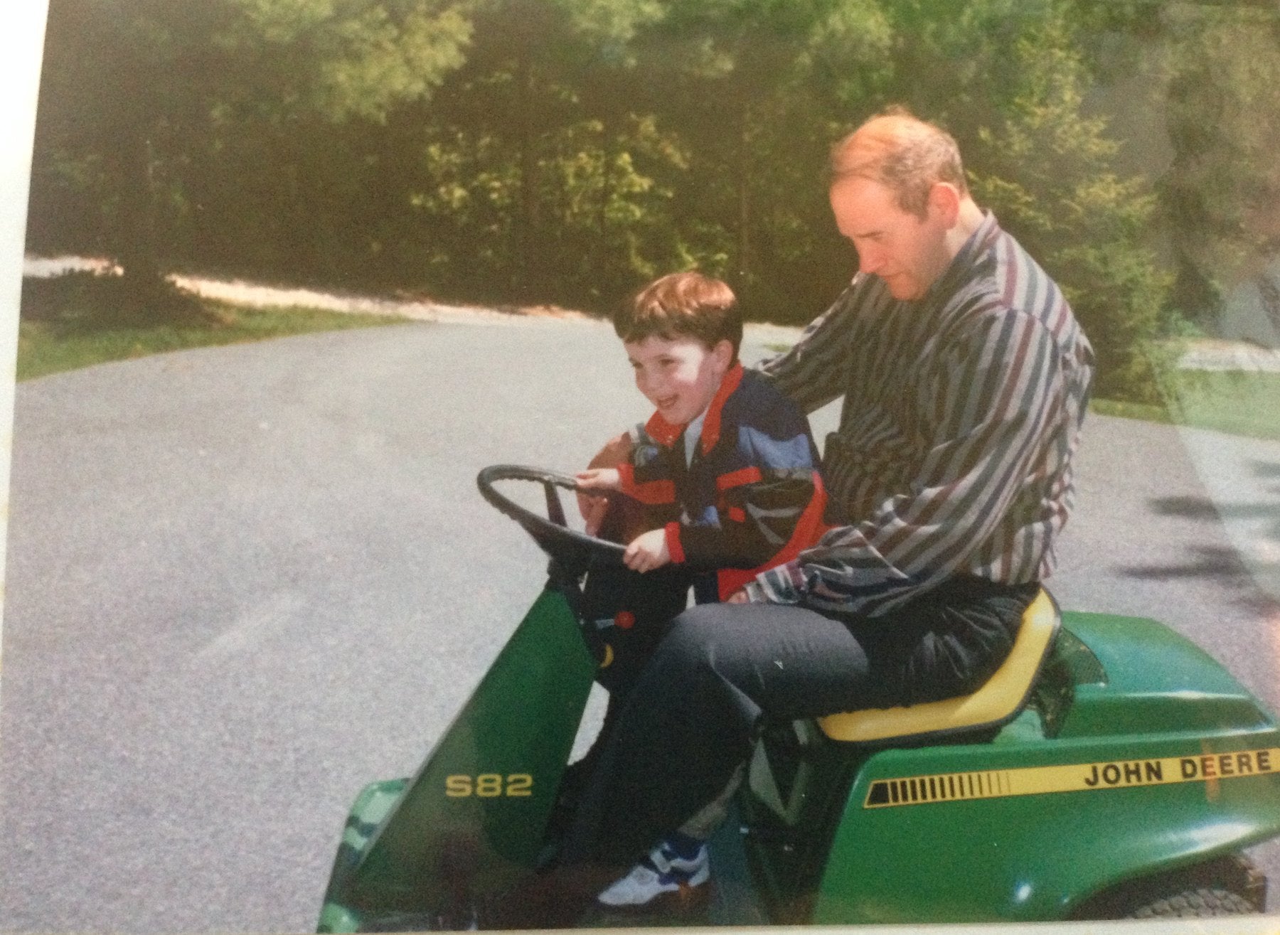 A young Mike Hoatson at the wheel of a green John Deere riding mower, sitting on the lap of his grandfather, Pop-Pop Donald Hoatson.